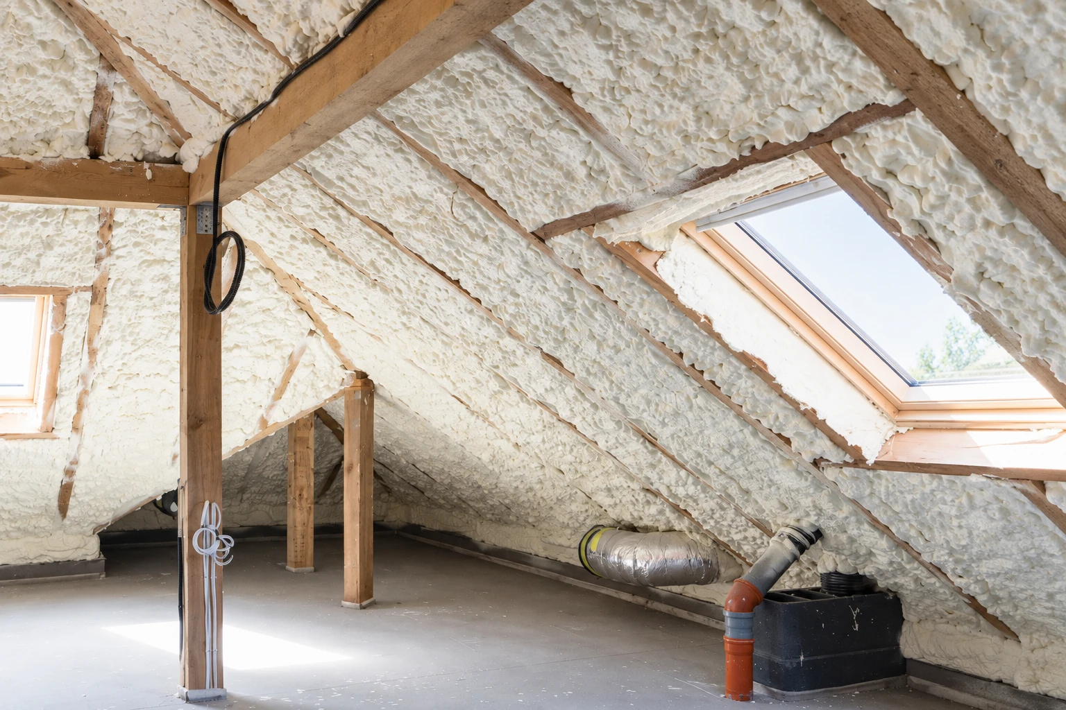 Attic rafters and roof slopes coated with white spray foam insulation, with skylights and ducts visible.