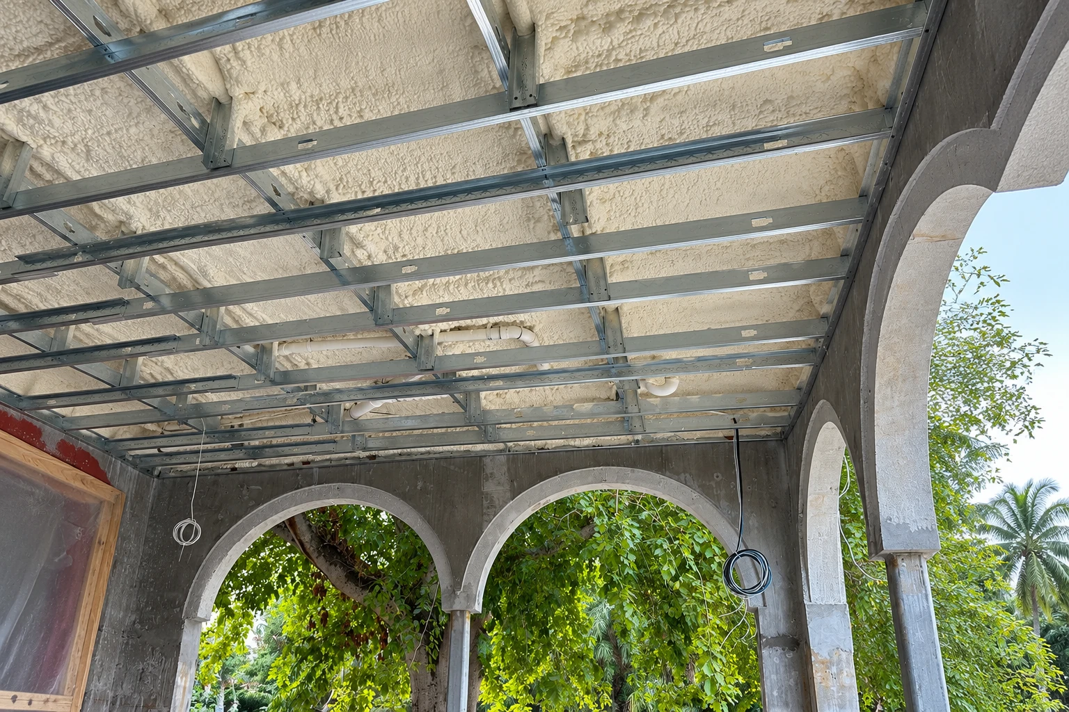 Spray foam coats the ceiling above a metal framing grid in an unfinished arched outdoor structure.