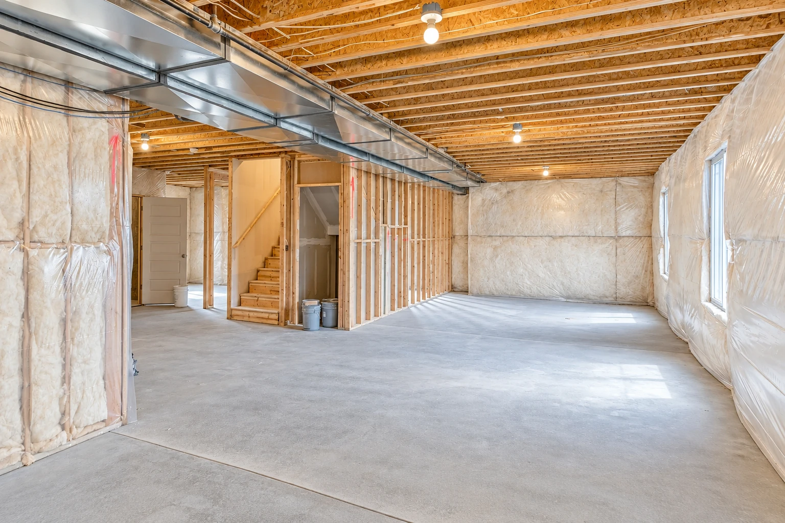 An unfinished basement has fiberglass insulation in its walls, exposed ceiling joists, metal ductwork, and a bare concrete floor.