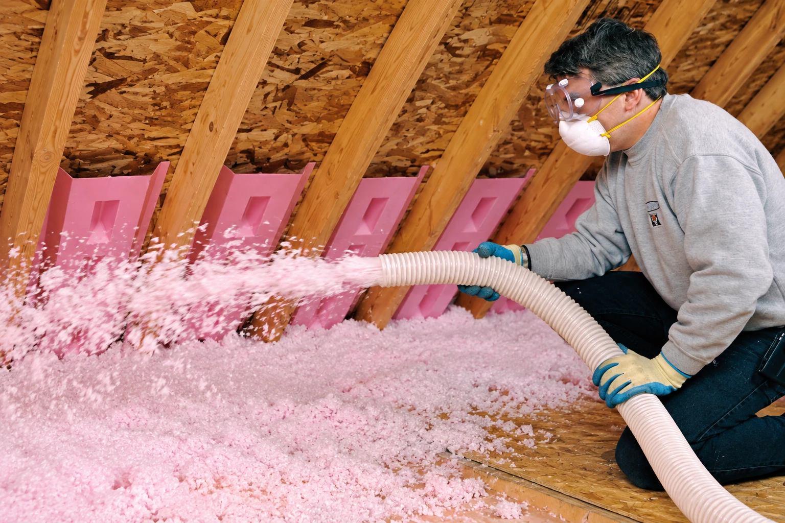 A worker in protective gear blows pink insulation into an attic between wooden rafters.