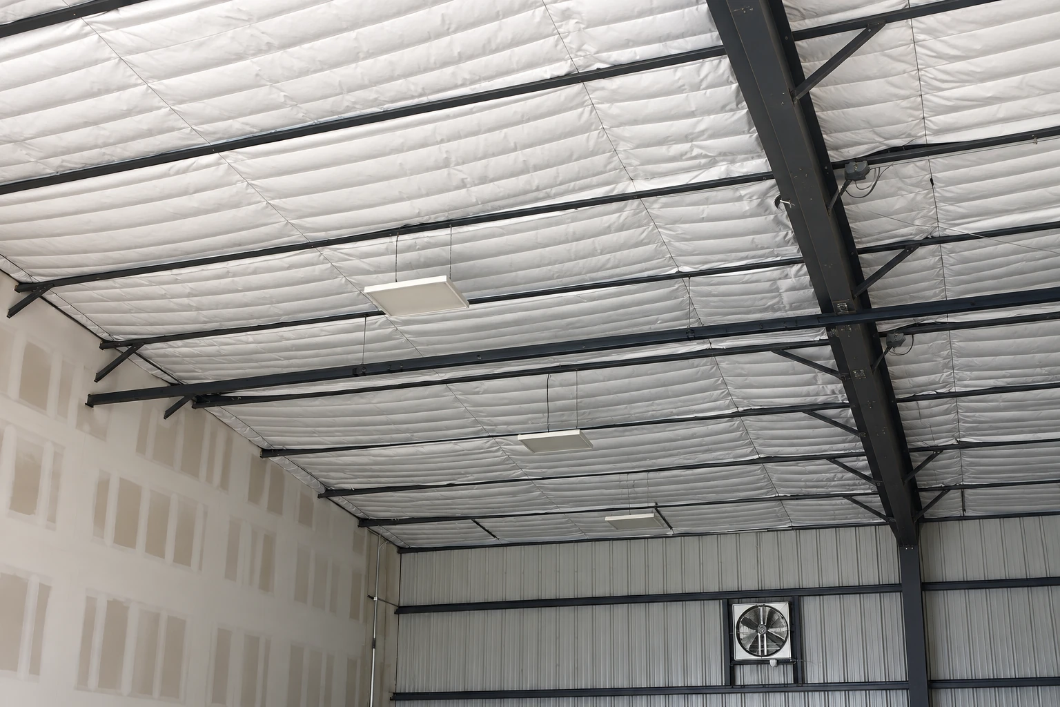 White insulation blankets the ceiling of a large metal-framed building with dark steel beams and a wall-mounted fan.