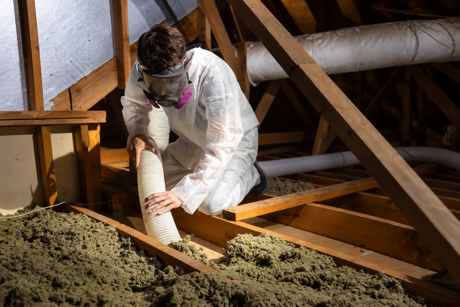 A worker wearing protective gear uses a hose to remove loose insulation from an attic between wooden joists.