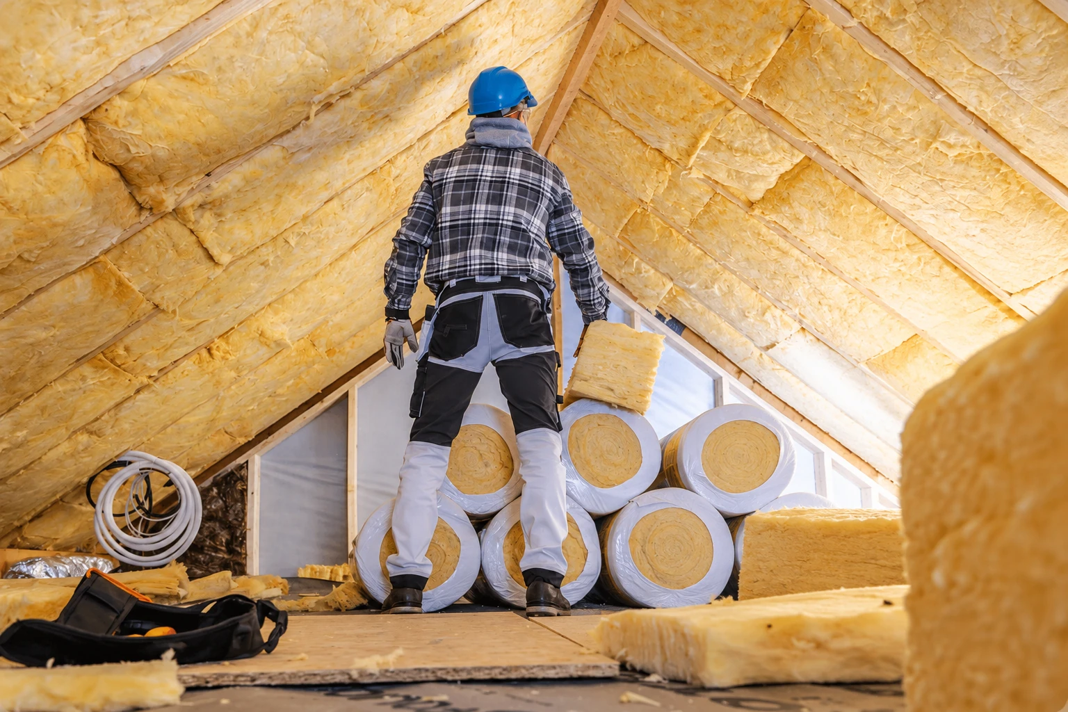 A worker in a blue hard hat stands in an attic lined with yellow fiberglass insulation, with rolls and loose batts nearby.