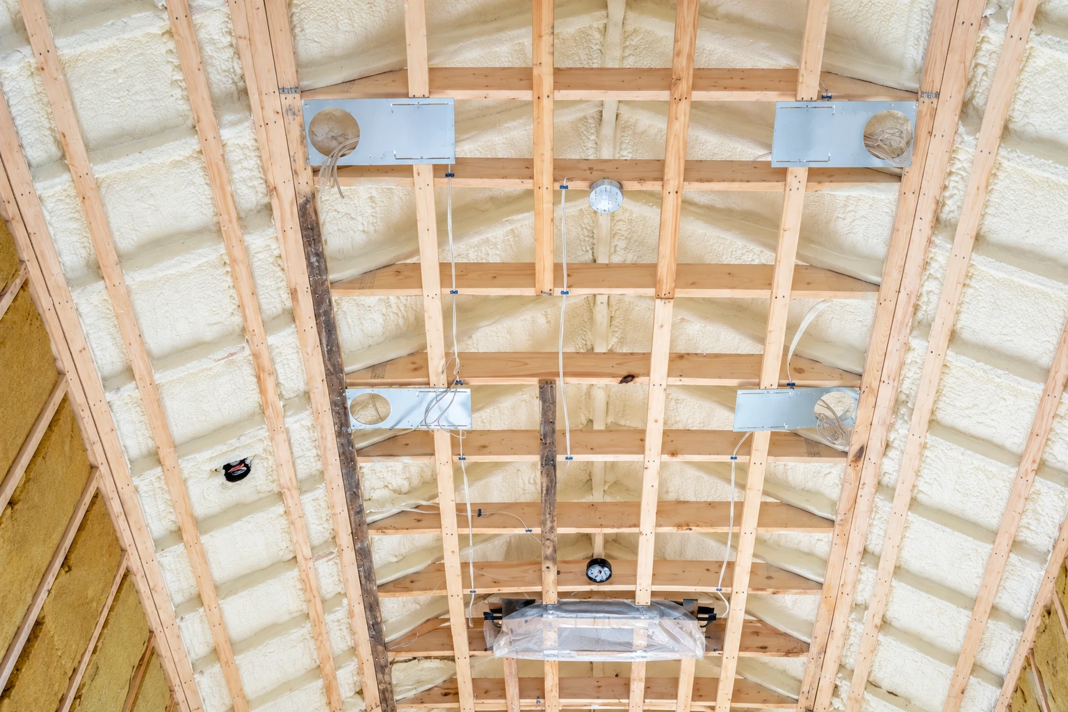 Spray foam insulation coats the roof between exposed wooden rafters, with metal boxes and wiring attached.