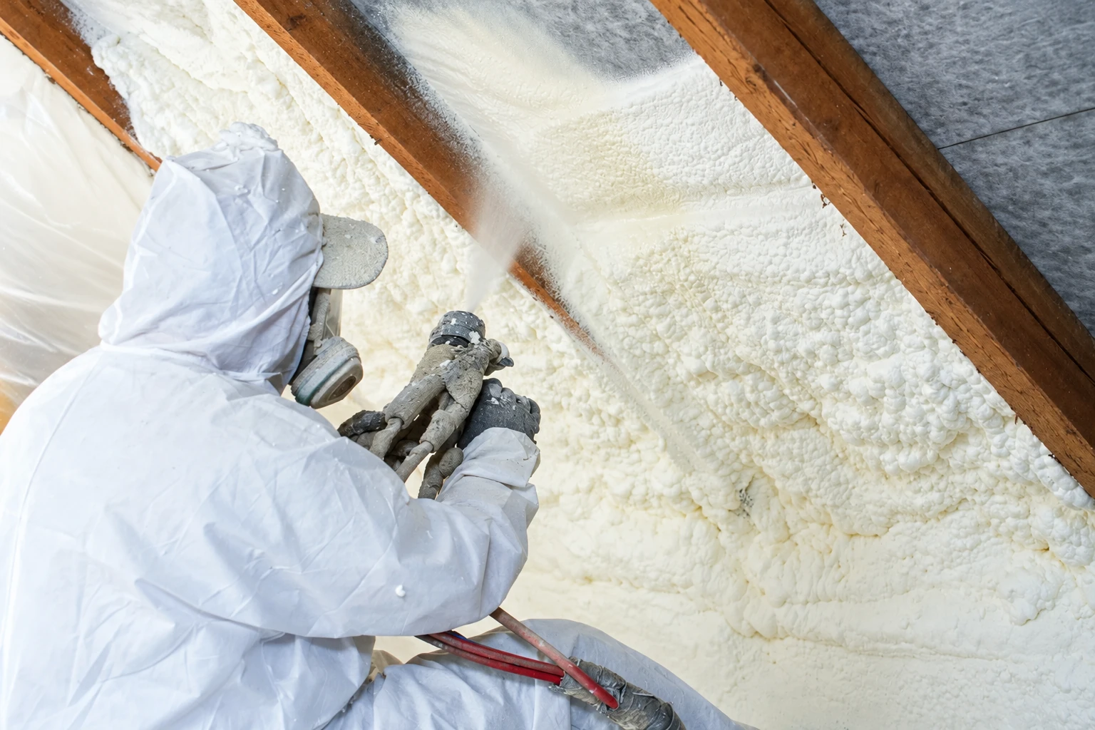 A worker in protective coveralls and a respirator sprays white foam insulation between exposed attic rafters.