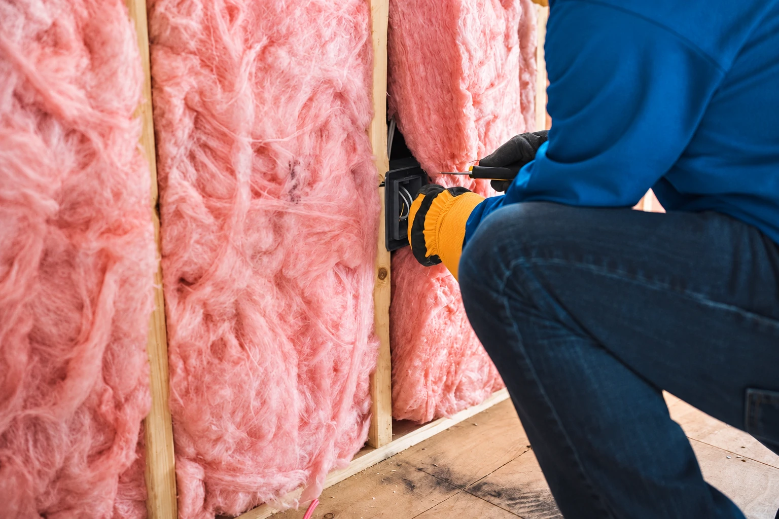 A worker in a blue jacket fits pink fiberglass insulation between wooden wall studs.